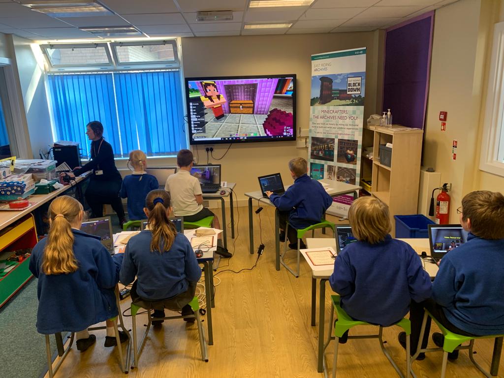 Photograph of a school classroom including 7 pupils sat at laptops and a facilitator directing the class via a large screen
